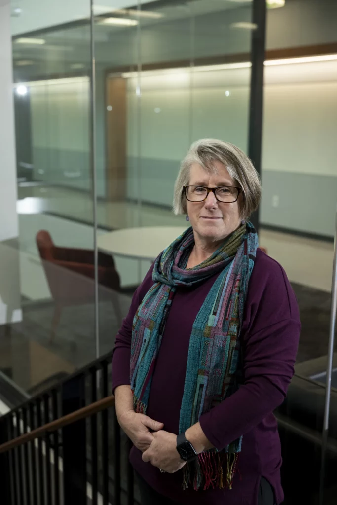 C. Robin Buell, wearing a purple sweater and patterned scarf, stands by a glass-walled hallway