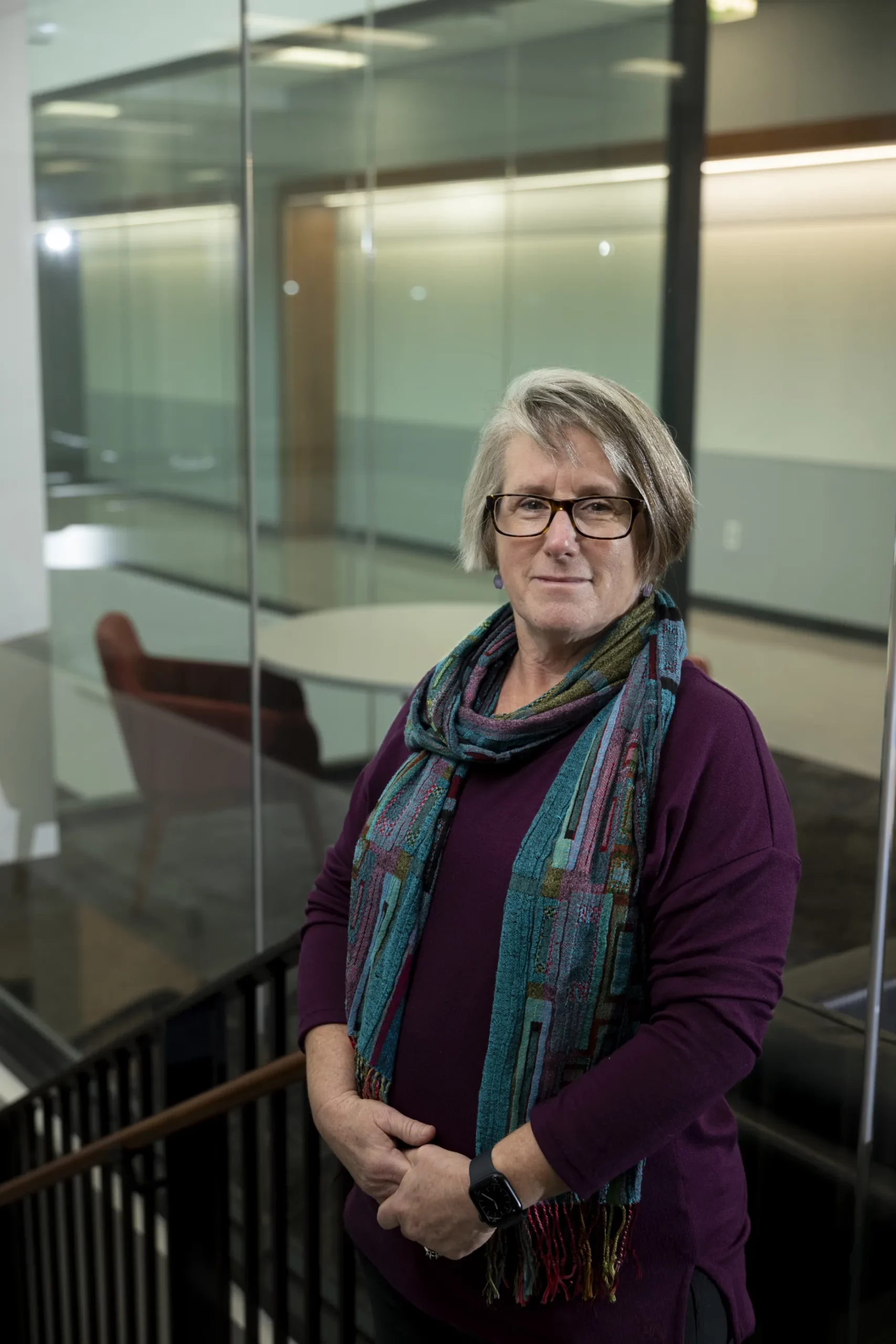 C. Robin Buell, wearing a purple sweater and patterned scarf, stands by a glass-walled hallway