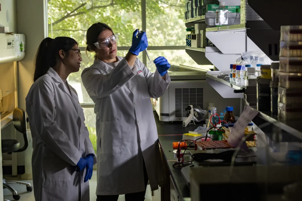 Two researchers in lab coats and safety gear examine a sample at a laboratory bench