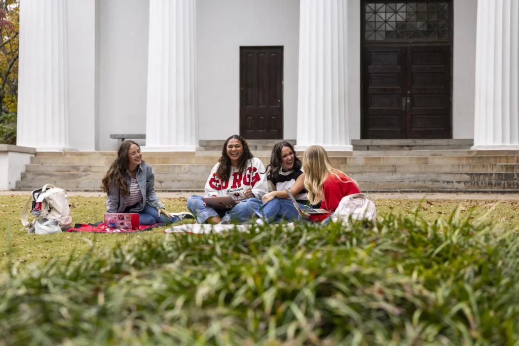 Four students laugh together on blankets on the lawn in front of a white-columned building