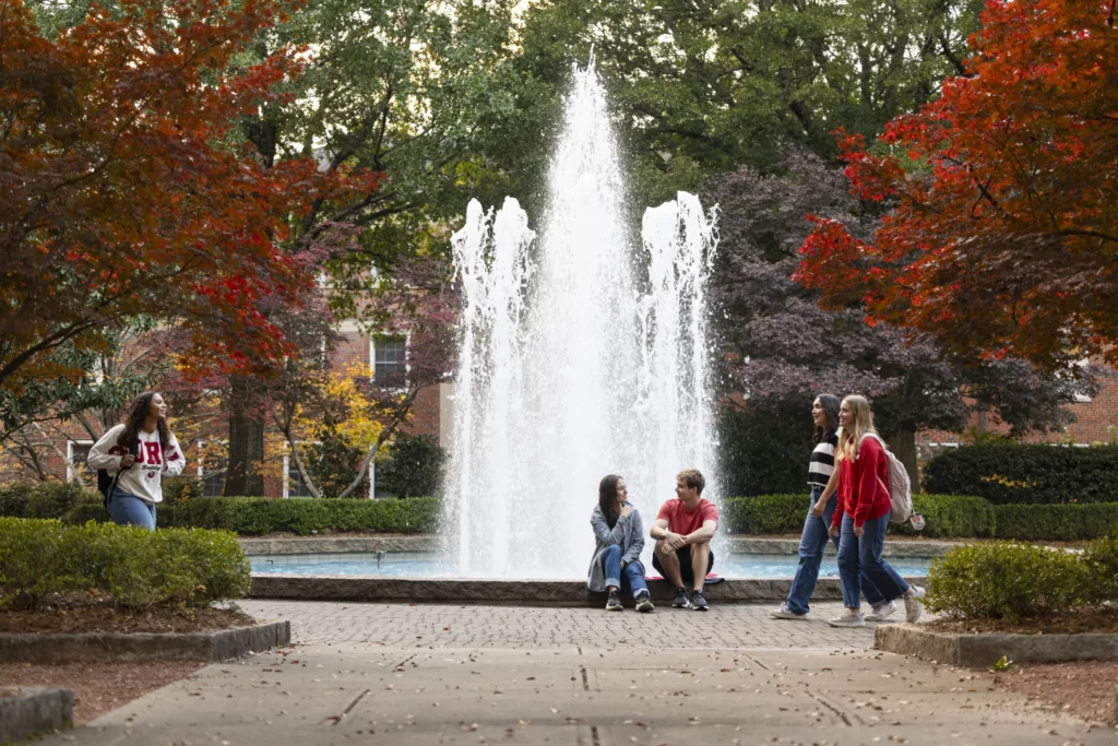 Students sit and walk near a tall fountain framed by red autumn trees on campus