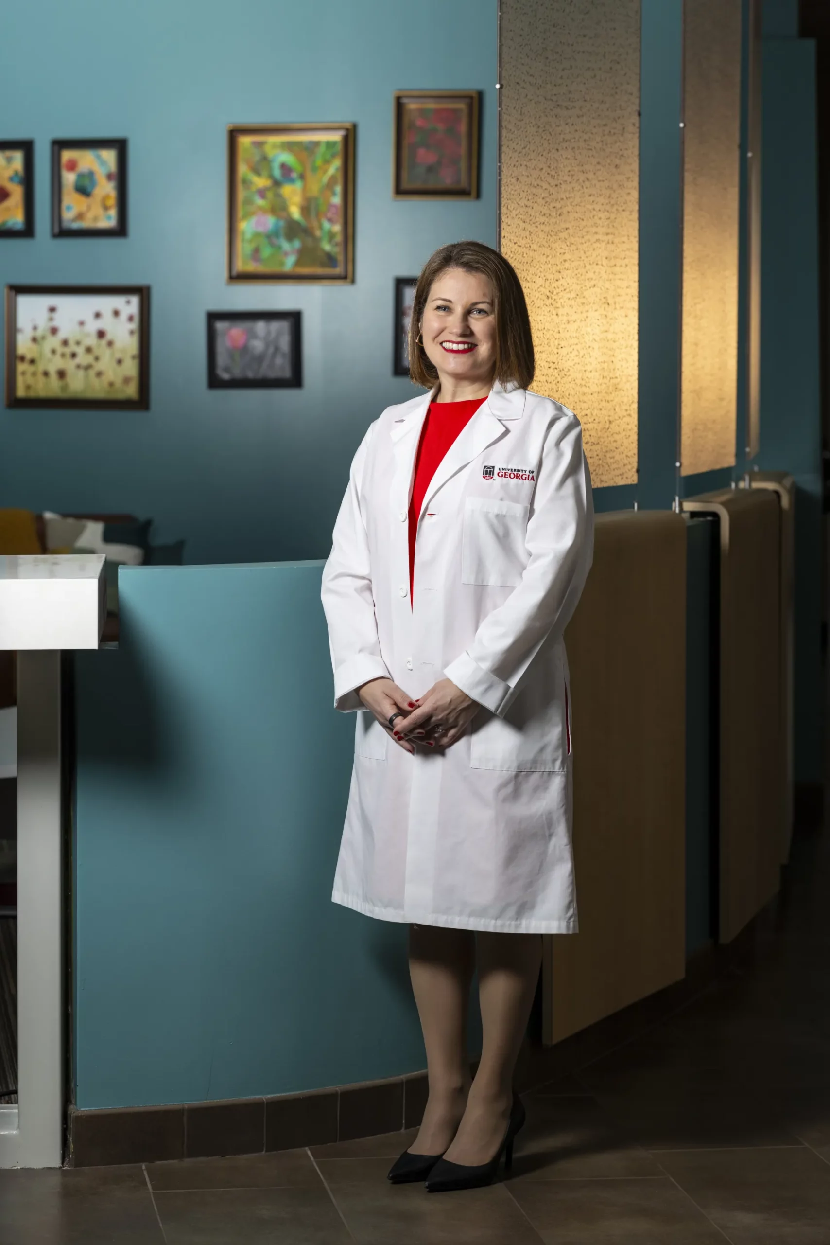 Carolyn Clevenger, in a white UGA lab coat, stands in a teal room decorated with framed paintings