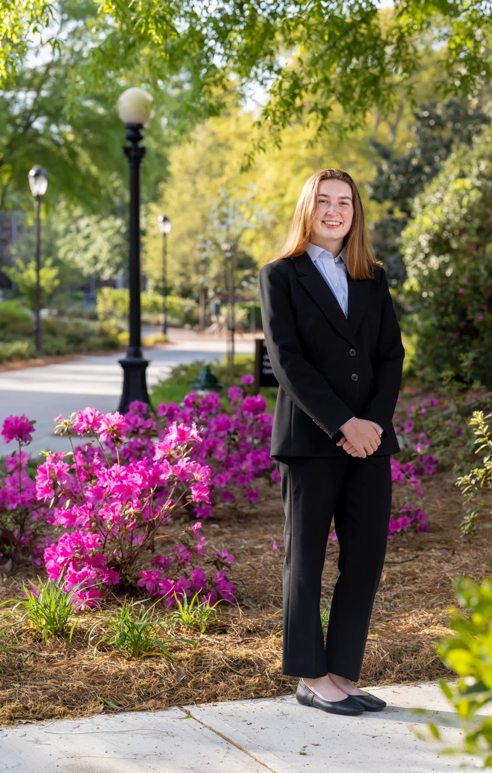 Finn Walsh, in a dark suit, stands beside pink azaleas along a lamp-lined campus walkway