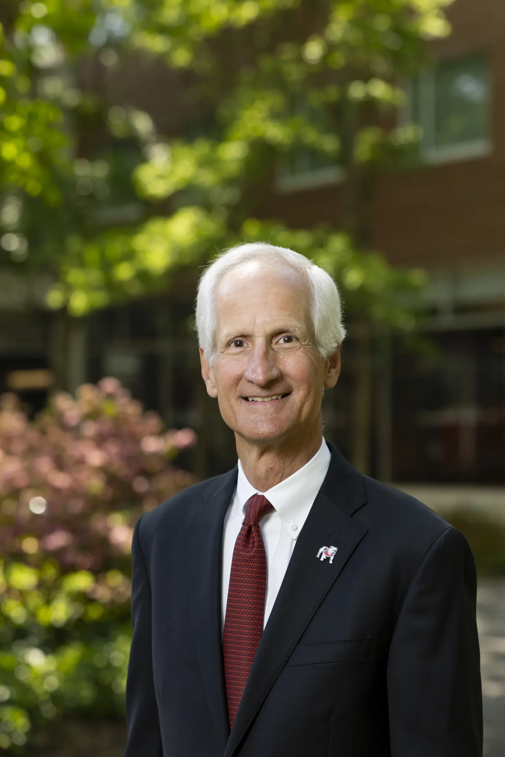 E. Howard Young, in a navy suit and red tie, stands outdoors beside flowering shrubs