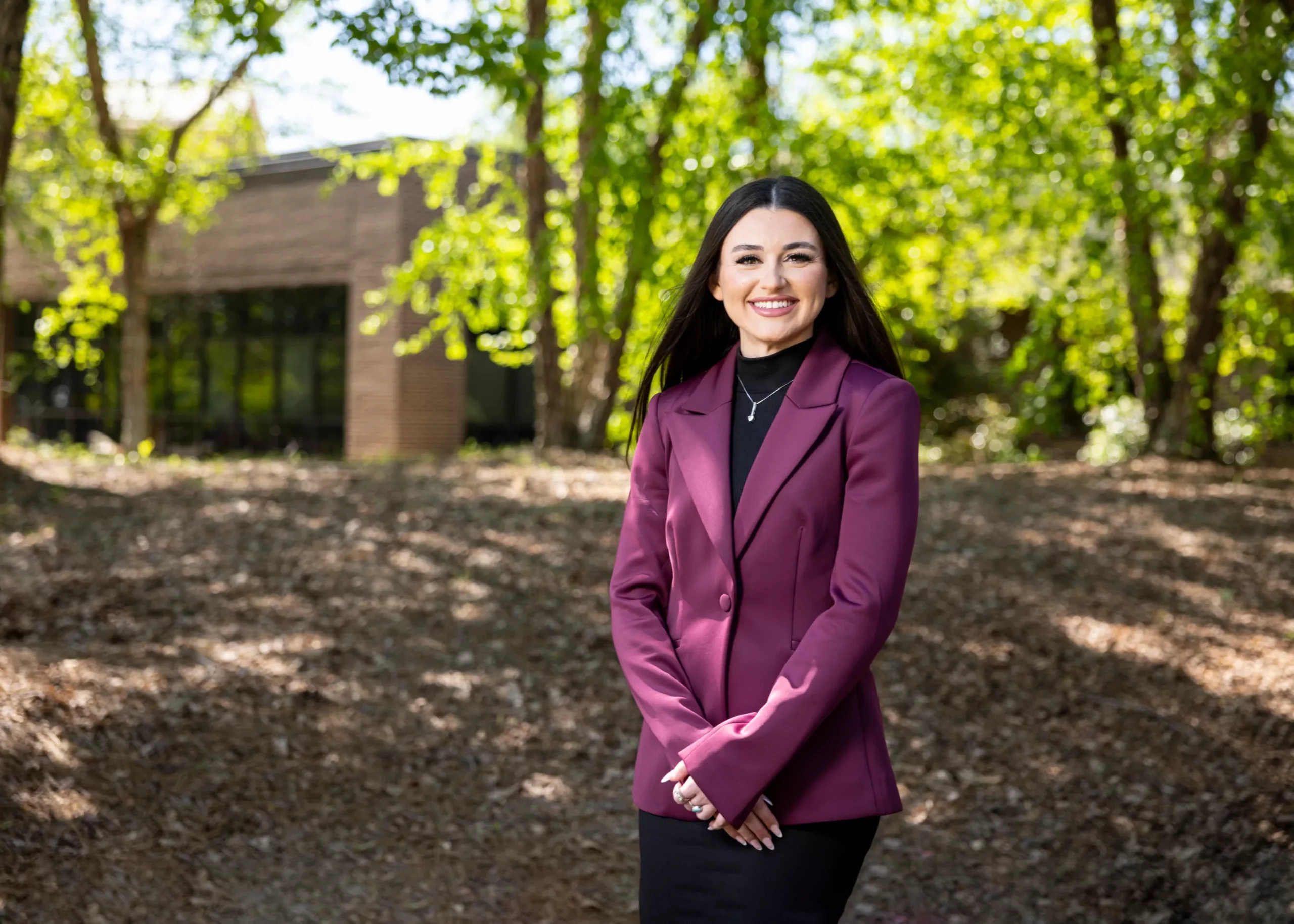 Ashley Galanti, in a plum blazer, stands in a sunlit wooded area on campus