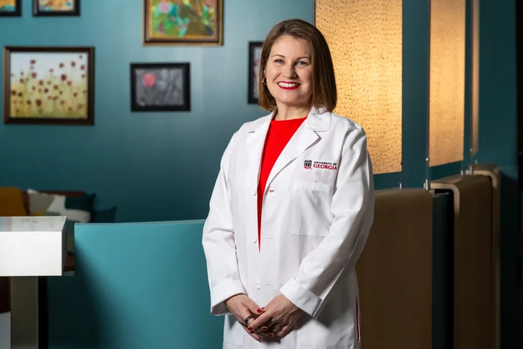 Carolyn Clevenger, in a white UGA lab coat, smiles in a teal room decorated with framed paintings