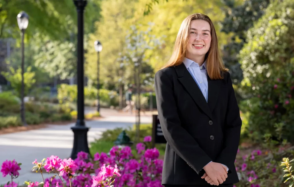 Finn Walsh, in a dark suit, smiles beside pink azaleas along a campus walkway